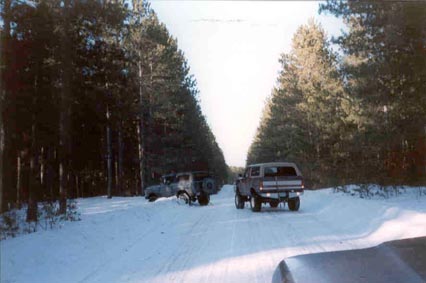 1991 Ford Bronco - Two SUVs travel down a snow-covered road flanked by tall pine trees.