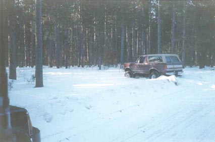1991 Ford Bronco - A red Ford Bronco with a camper shell is partially buried in snow in a wooded area.
