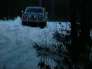 1991 Ford Bronco - A dark-colored SUV navigates a snow-covered road through a wooded area, kicking up snow as it moves.