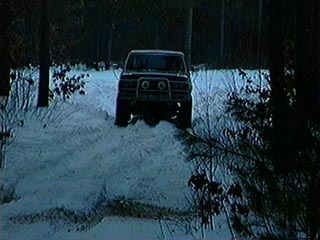 1991 Ford Bronco - A dark colored SUV navigates a snow-covered trail through a wooded area.