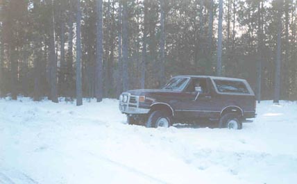 1991 Ford Bronco - A dark red Ford Bronco is partially submerged in deep snow, surrounded by a dense forest.