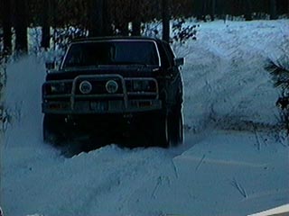 1991 Ford Bronco - A black SUV navigates a snowy path, kicking up snow as it drives forward.