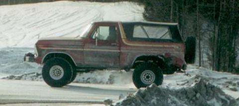 1991 Ford Bronco - A red 1978 Ford Bronco with a black soft top drives through snow, its lifted suspension and large tires evident.