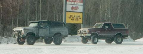 1991 Ford Bronco - Two Ford Broncos, one maroon and one gray, are parked in a snowy landscape in front of a "Hot Stuff Pizza" sign.