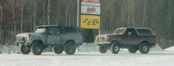 Gray and maroon lifted Ford Broncos parked on snow in front of a pizza restaurant sign