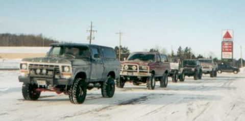 1991 Ford Bronco - Several SUVs and trucks are lined up on a snowy road, with a gas station sign in the background.