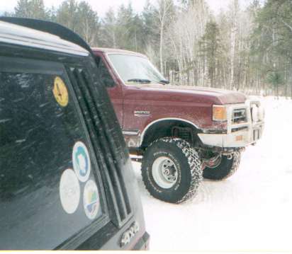 1991 Ford Bronco - A red 1991 Ford Bronco is parked in a snowy, wooded area, partially obscured by another vehicle.