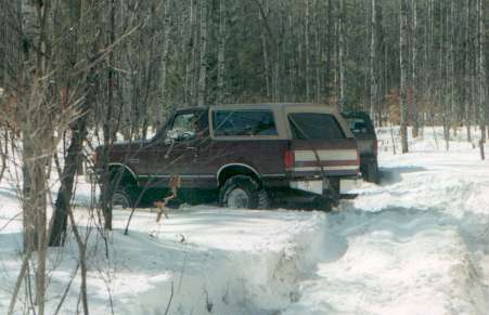 1991 Ford Bronco - A maroon 1991 Ford Bronco is partially buried in snow, with another vehicle visible behind it in a wooded area.
