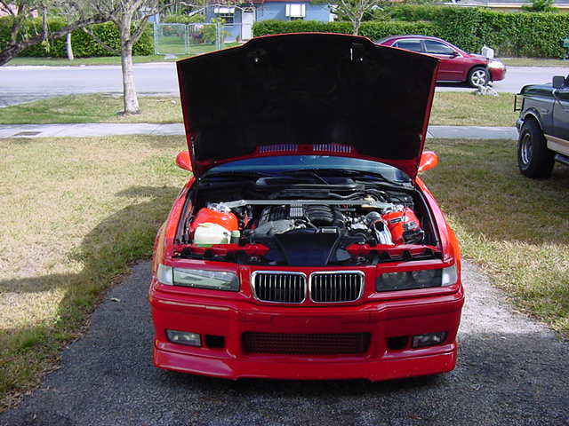 Red BMW E36 coupe photographed head-on with hood raised showing engine bay