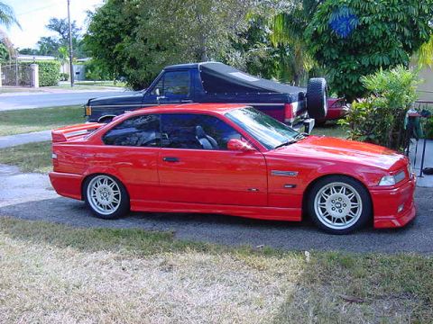 1995 BMW M3 - A red 1995 BMW M3 coupe is parked in front of a blue truck. The M3 features distinctive wheels and a rear spoiler.
