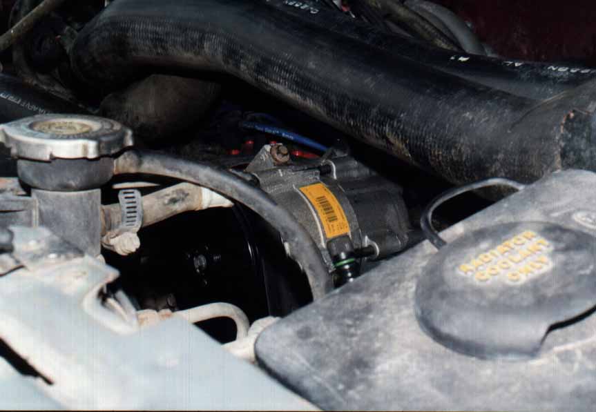 Engine bay close-up of A/C compressor with yellow label near coolant reservoir and radiator cap