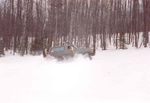 1995 Ford Bronco - A green Ford Bronco navigates a snowy forest, kicking up snow as it turns.