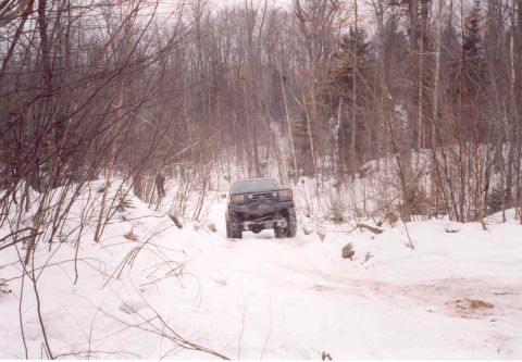 1995 Ford Bronco - A dark SUV navigates a snow-covered path through a wooded area, suggesting a 1990s Ford Bronco.