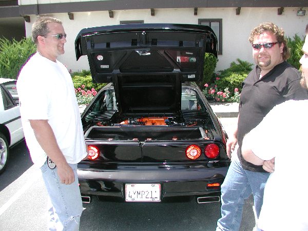 1986 Pontiac Fiero, rear view, trunk open showing orange engine. Two men stand nearby