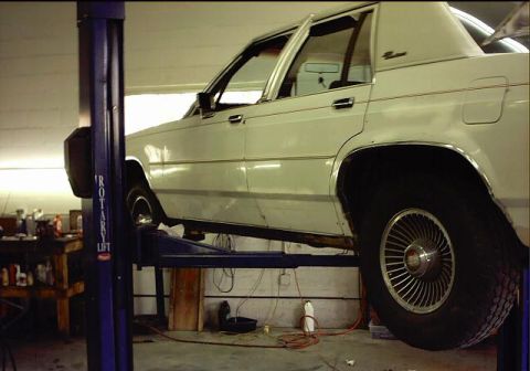 1985 Ford Crown Victoria LTD - A white sedan is raised on a car lift in a garage, showing its side profile and underside.