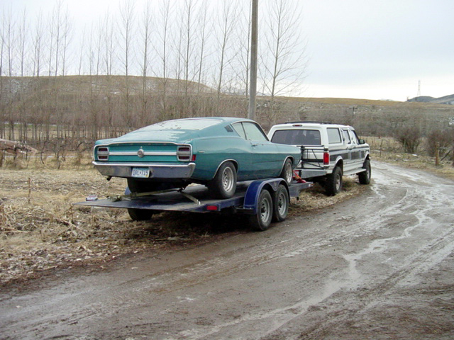 Teal fastback coupe on trailer towed by white pickup on muddy road