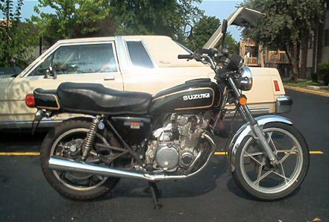 1985 Ford LTD Crown Victoria - A Suzuki motorcycle is parked in front of a car with its hood open.