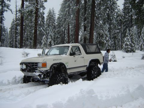 1991 Ford Bronco - A modified white Ford Bronco navigates deep snow in a wooded area.