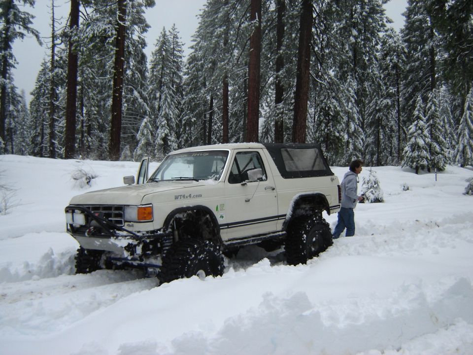 White lifted Ford Bronco on big tires in deep snow among pine trees