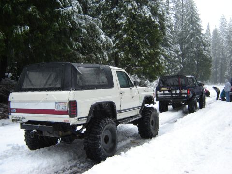 1991 Ford Bronco - Two modified SUVs navigate a snow-covered road in a wooded area.