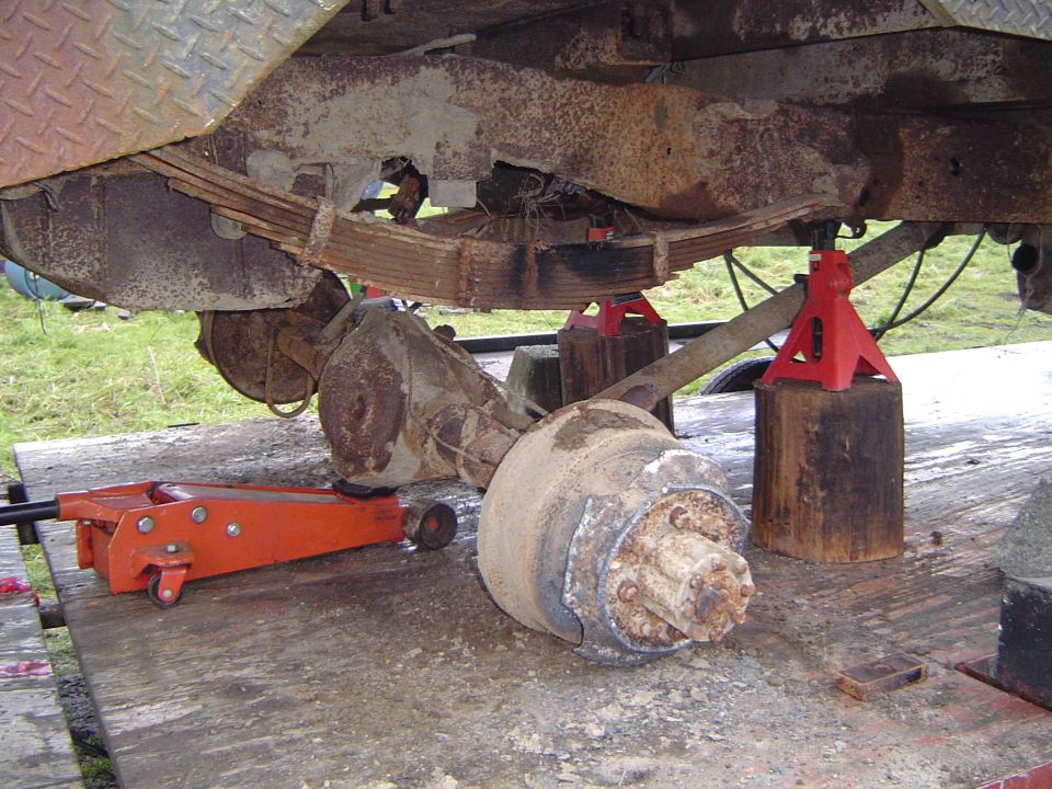 Rear leaf-spring axle on jack stands with hub exposed, floor jack under the differential, truck up on planks