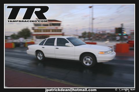 1997 Ford Crown Victoria - A white 1997 Ford Crown Victoria is captured in motion on a race track, with a building and spectators visible in the background.