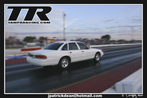 1997 Ford Crown Victoria - A white 1997 Ford Crown Victoria accelerates down a drag strip under a cloudy sky.