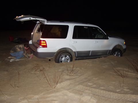 1979 Ford Bronco - A person attempts to free a white SUV that is stuck in the sand at night, with the rear hatch open.