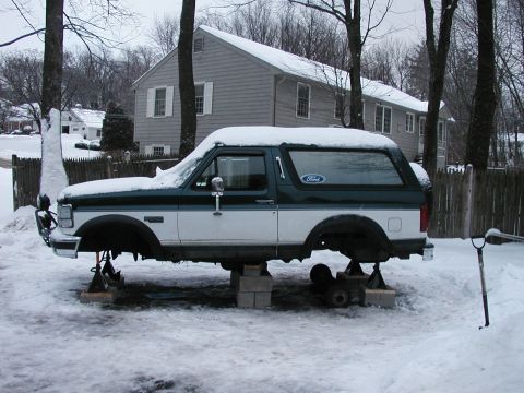 1994 Ford Bronco - A two-tone Ford Bronco sits on blocks and jack stands in the snow, missing its wheels.