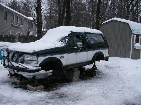1994 Ford Bronco - A two-tone Ford Bronco sits on jack stands in a snowy yard, its wheels removed for maintenance.