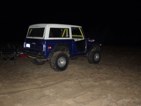 1979 Ford Bronco - A blue and white 1979 Ford Bronco with a roll cage sits on a sandy area at night.