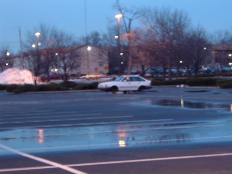 1995 Mercury Grand Marquis - A white sedan sits in a wet parking lot as dusk settles, with streetlights illuminating the scene.