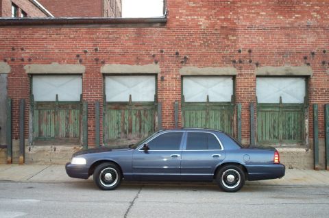 1998 Ford Crown Victoria - A blue 1998 Ford Crown Victoria is parked on the street in front of a brick building with weathered green doors.