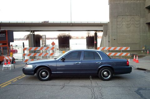 1998 Ford Crown Victoria - A dark blue 1998 Ford Crown Victoria is parked on a street with flood barriers and cones in the background.