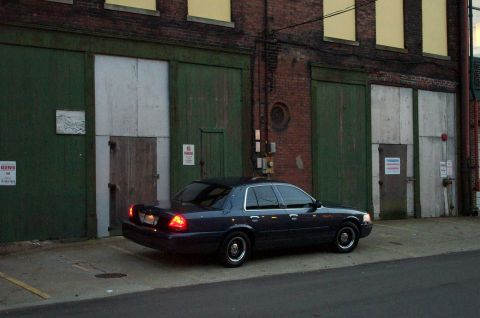1998 Ford Crown Victoria - A dark sedan is parked in front of an old industrial building with large bay doors.