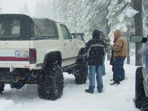 1986 Ford Bronco - Several people gather around a lifted off-road vehicle in a snowy forest environment.