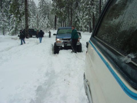1986 Ford Bronco - A blue pickup truck is stuck in the snow in a wooded area, with several people around.