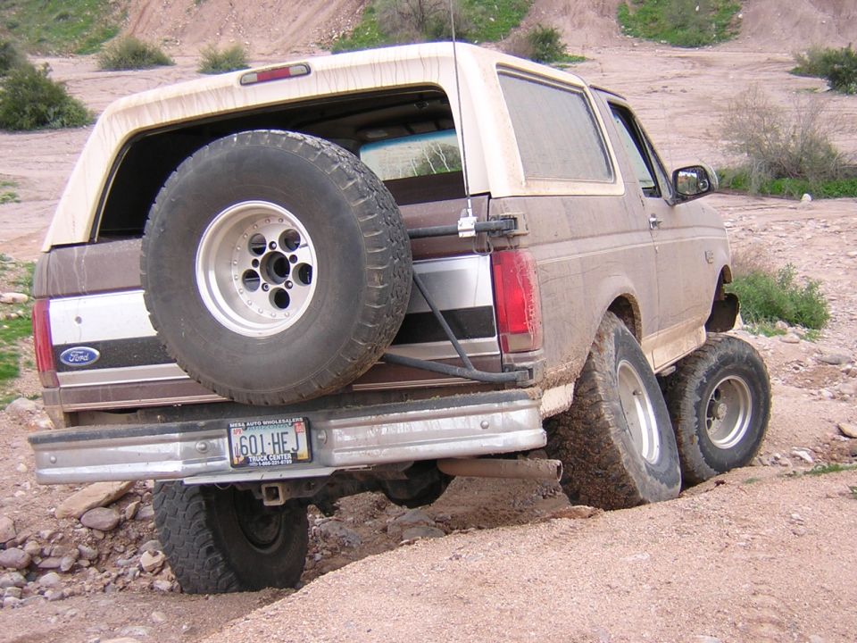 Rear of muddy tan Ford Bronco flexing over a dirt ledge, tire carrier and license plate visible
