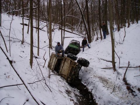 1992 Ford Bronco - Two off-road vehicles struggle in the snow on a wooded slope, with one vehicle overturned.