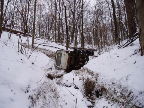 1992 Ford Bronco - A white truck lies on its side in a snow-filled ditch amidst a wooded area.
