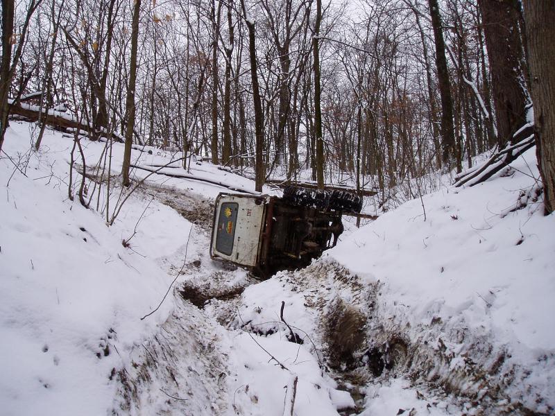 White truck overturned in a snowy ditch with trees in the background
