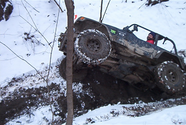 Off-road vehicle with large tires climbing a muddy, snowy hill. Driver visible