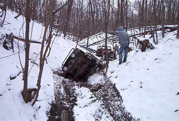 Two off-road vehicles are stuck in a steep, snowy ravine with trees all around. One vehicle is tilted on its side