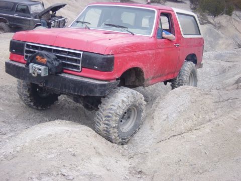 1983 Ford Bronco - This red SUV is navigating a rocky off-road trail, while another vehicle is being worked on in the background.