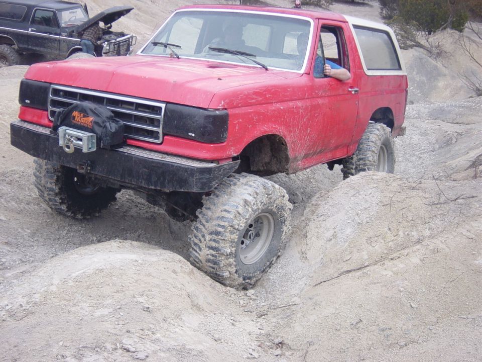 Red 1990s Ford Bronco with winch flexing over rock and dirt obstacle off-road