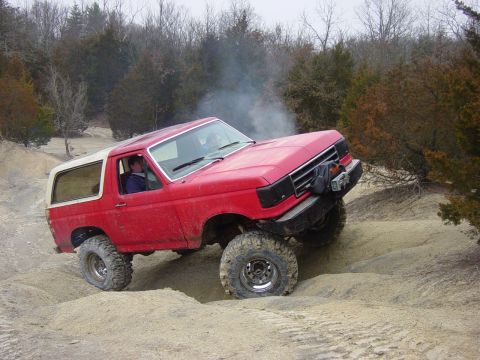 1983 Ford Bronco - This red SUV navigates a challenging dirt terrain, with visible smoke indicating engine strain.