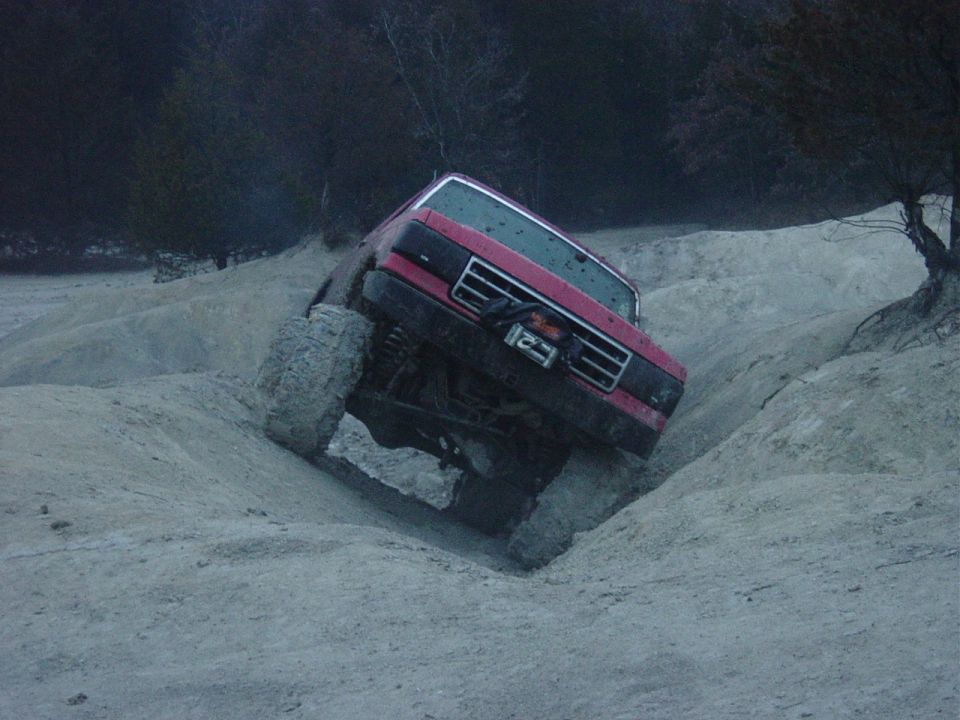 Red lifted Ford Bronco tilted steeply climbing out of a sandy gully at dusk