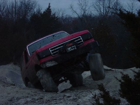 1983 Ford Bronco - A red SUV is shown tilted on a dirt path, with its wheels off the ground.