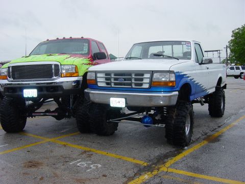 1983 Ford Bronco - White Ford F-150 pickup truck with blue flame graphics and lifted suspension, parked next to another lifted truck.