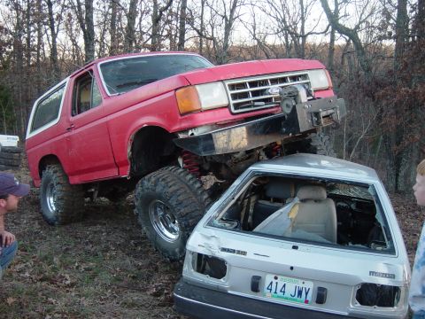 1983 Ford Bronco - Red Ford Bronco is on top of a wrecked silver Mazda. The Bronco appears to be modified for off-roading.